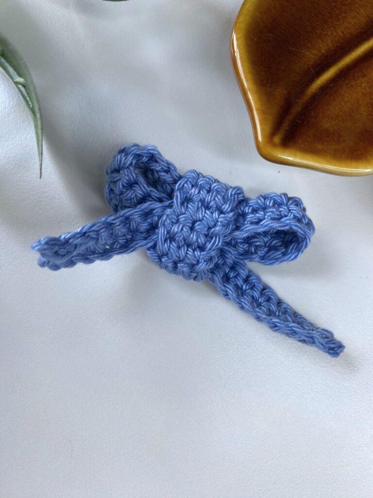 A crochet Christmas bow decoration is on a white surface with a brown leaf shaped dish above
