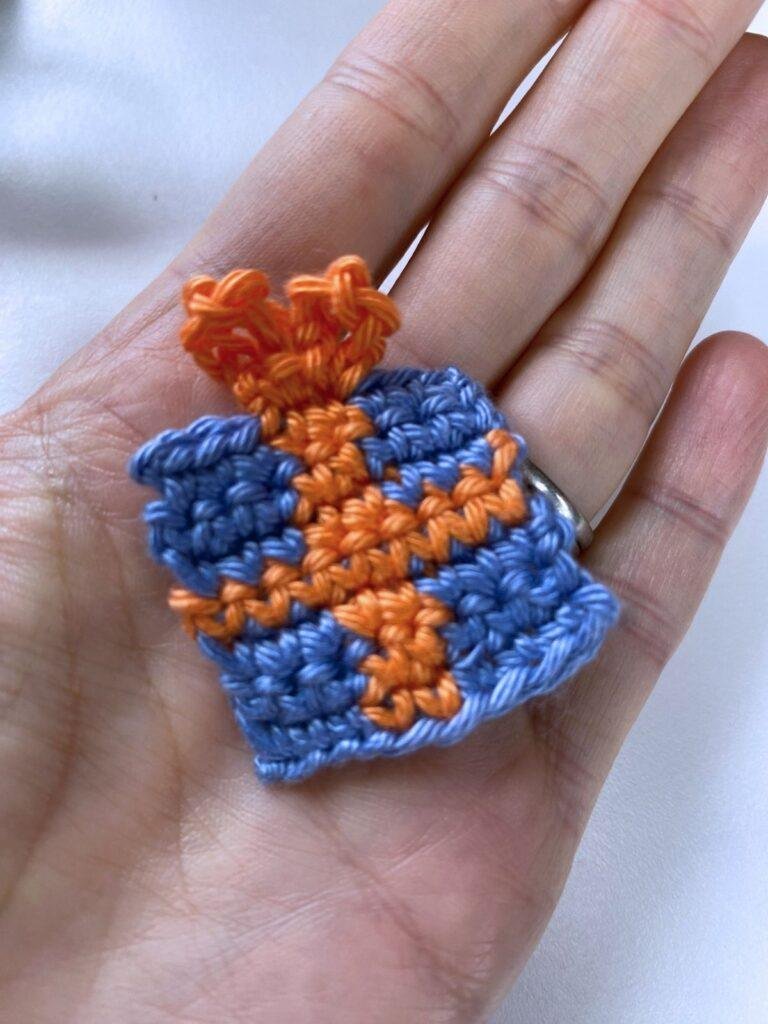 A hand holds a small crochet Christmas present motif in blue with orange crocheted ribbon