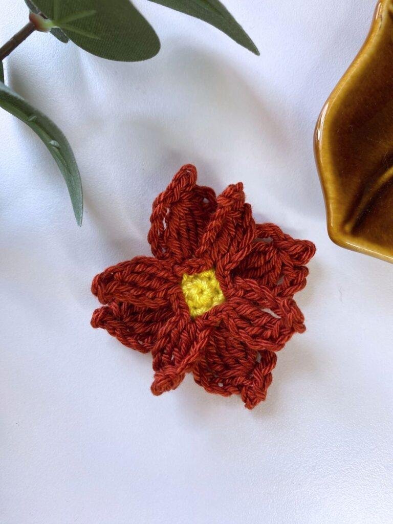 A crochet poinsettia flower decoration is on a white surface with greenery and a brown dish edge showing