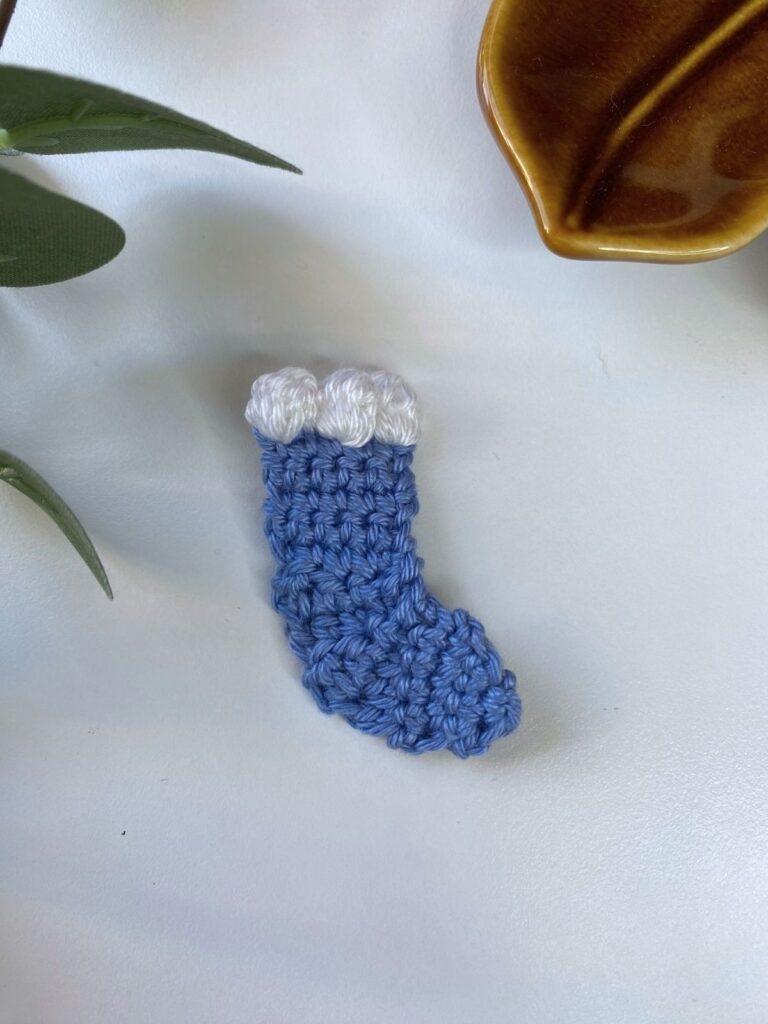 A mini blue crochet stocking ornament is on a white surface with leaves poking in from the edge