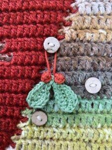 A small crochet mistletoe hangs on a button on a crochet advent calendar wall hanging