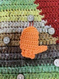 An orange crochet mitten ornament hangs on a button on a crochet Christmas tree picture