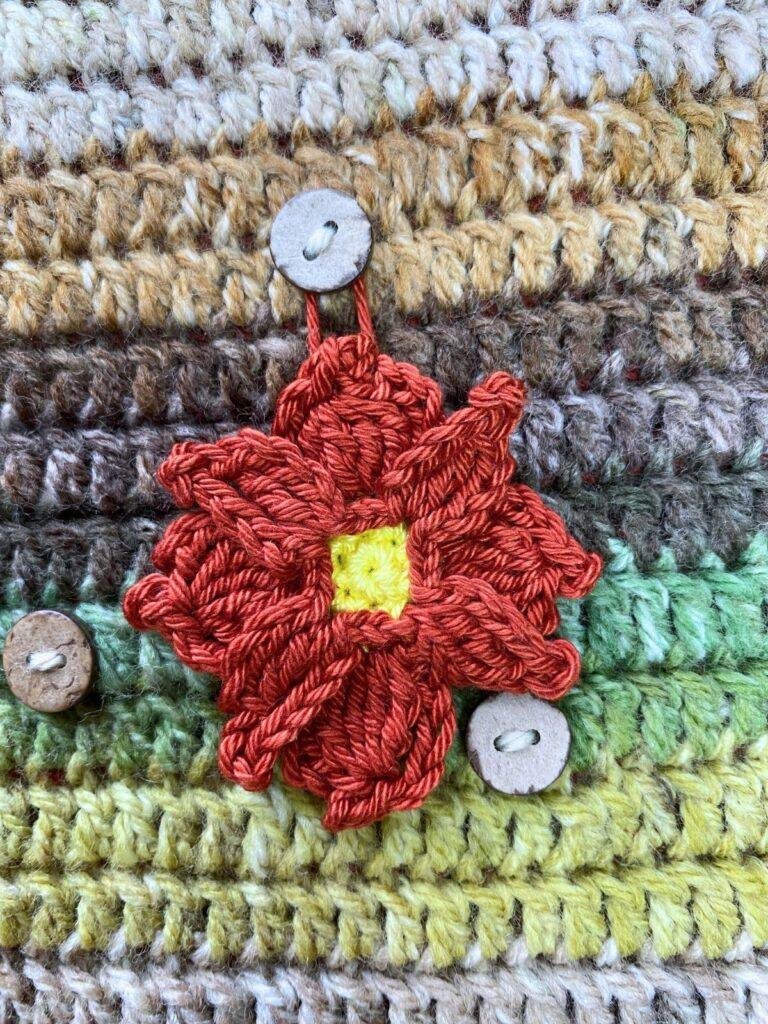 A crochet poinsettia with a yellow centre and red petals hangs on a green and brown crochet background