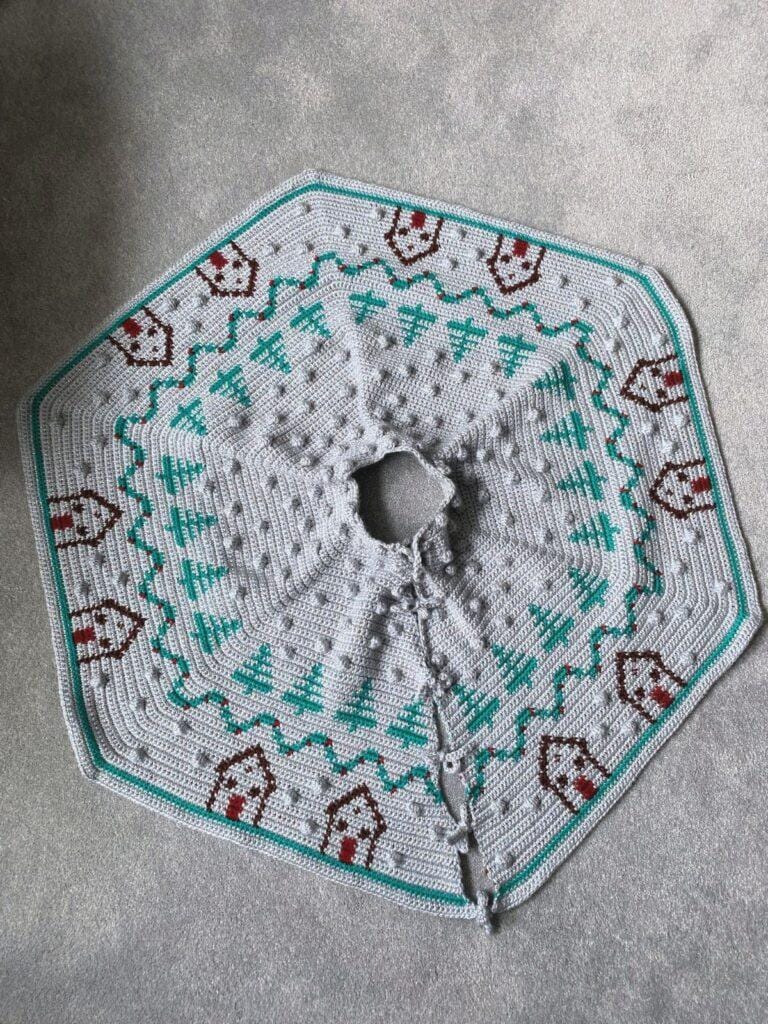 A hexagonal crochet silver Christmas tree skirt  is laid out on the floor. It features trees, and little brown huts
