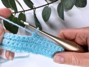 2 hands holding a gold crochet hook and yarn making a blue crochet fabric