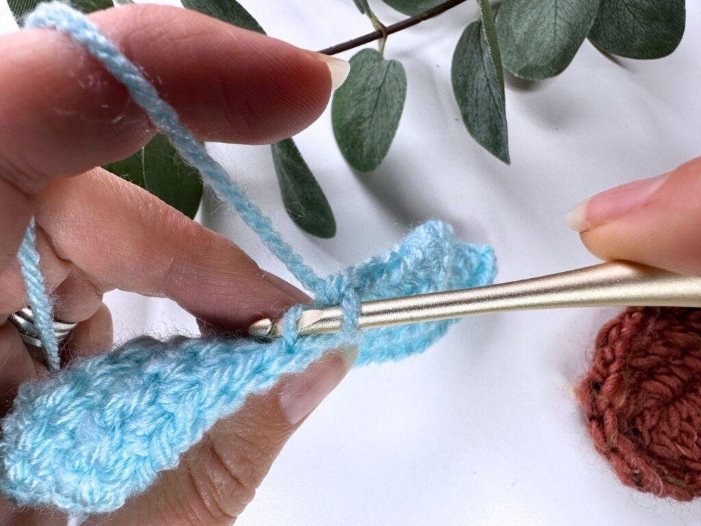 A persons hands showing the back loop of a stitch with a gold crochet hook in a piece of blue crocheted fabric