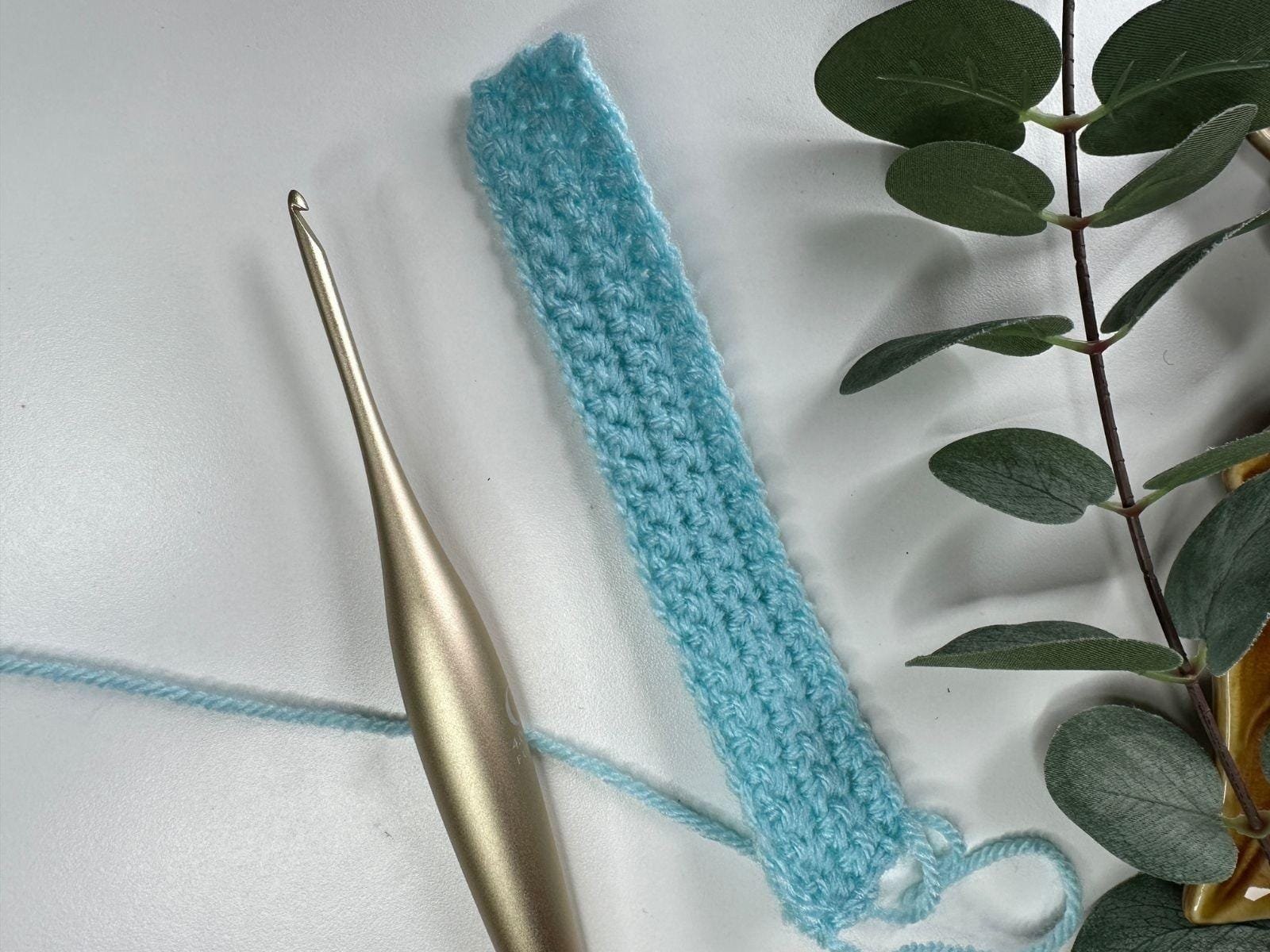 A white background with a blue swatch of crochet, a gold crochet hook and some greenery