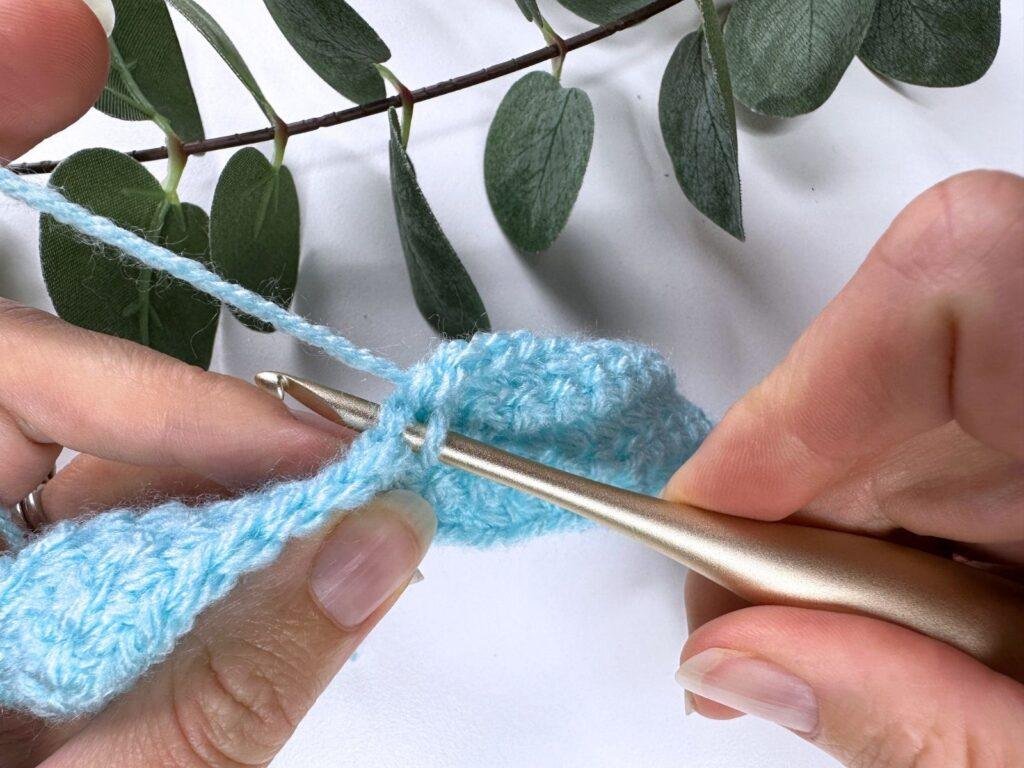 A person inserts a gold hook into a blue crochet piece of fabric to start making a stitch