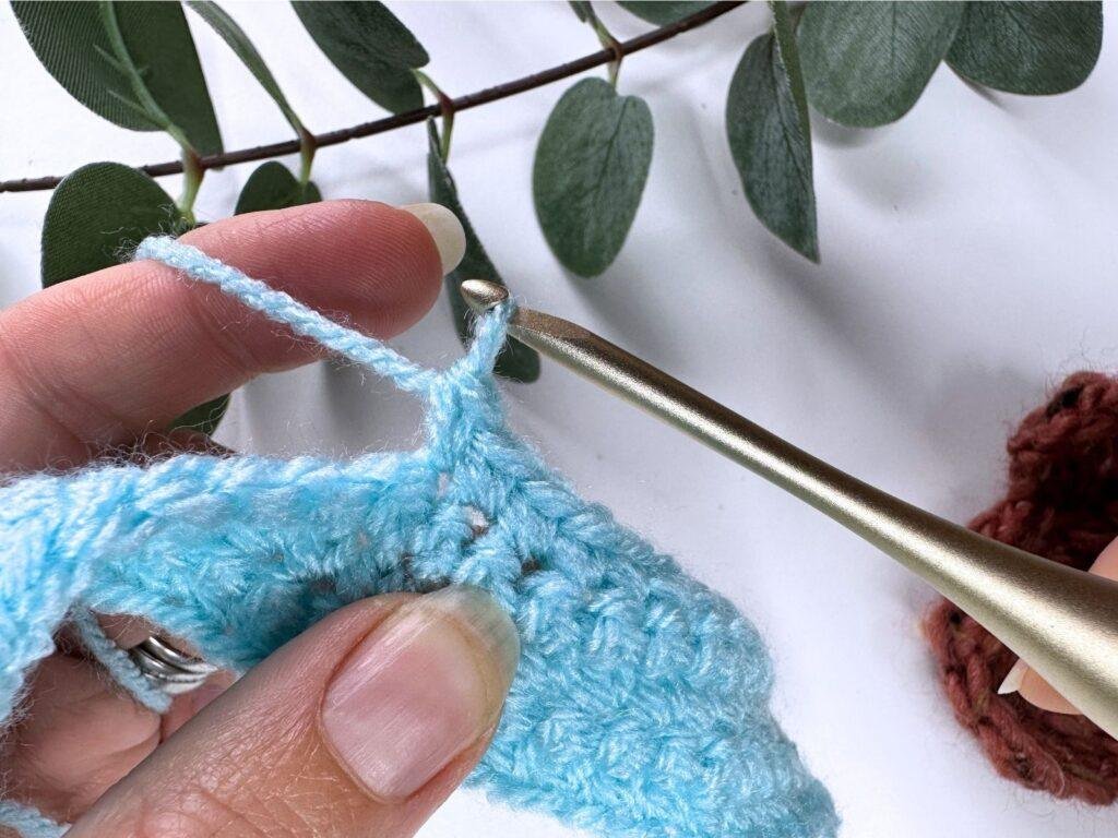 A person has just completed a single crochet stitch with blue yarn and a gold hook