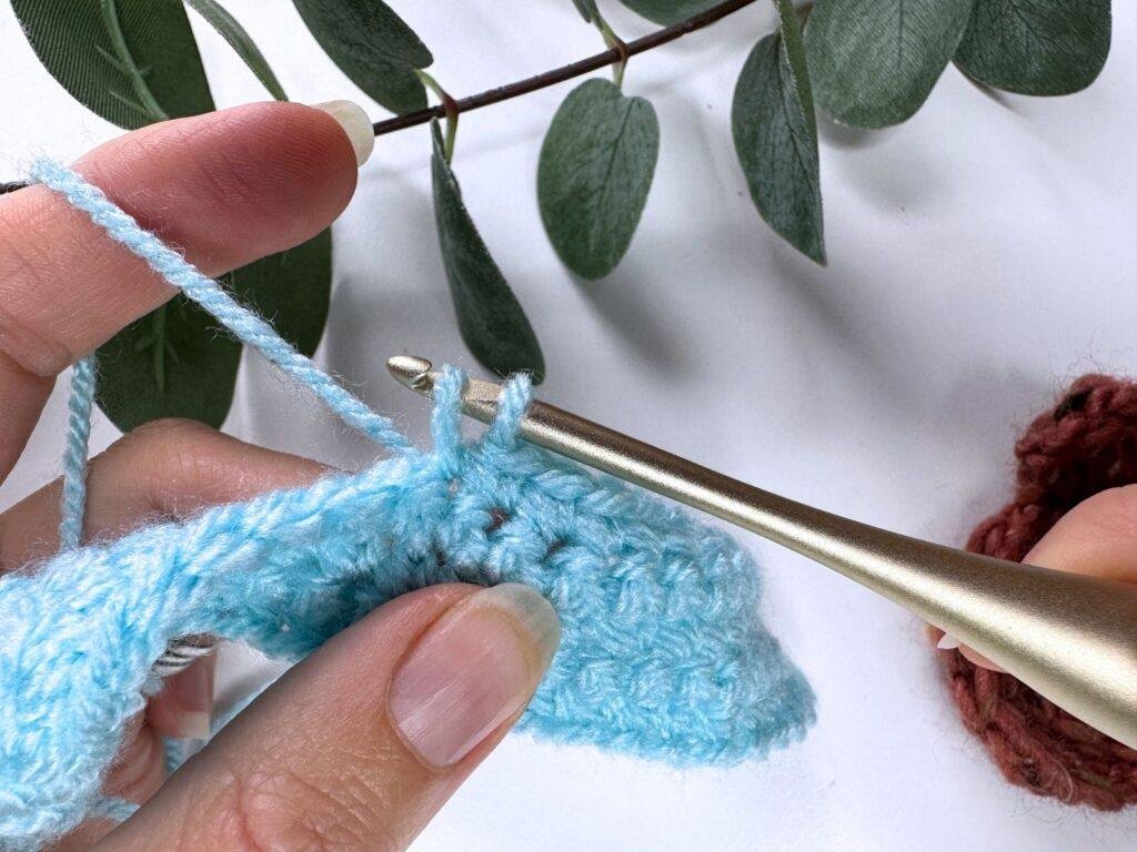 2 hands with blue yarn and a gold hook are demonstrating how to make a single crochet stitch