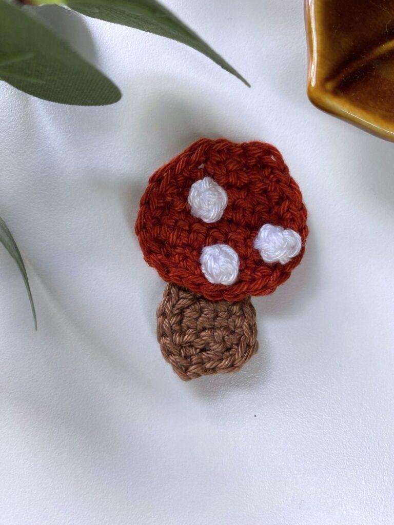 A toadstool crochet ornament is on a white table with greenery above and the edge of a brown dish