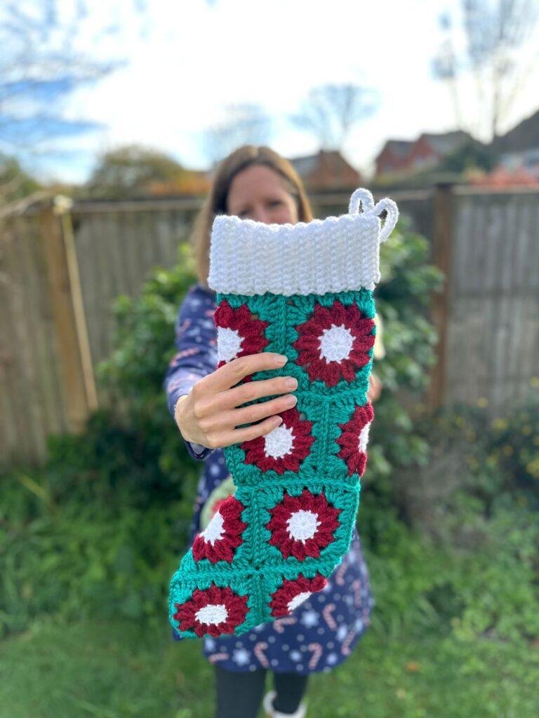A person stands in a garden and holds out a white, red and green crochet stocking in front of her