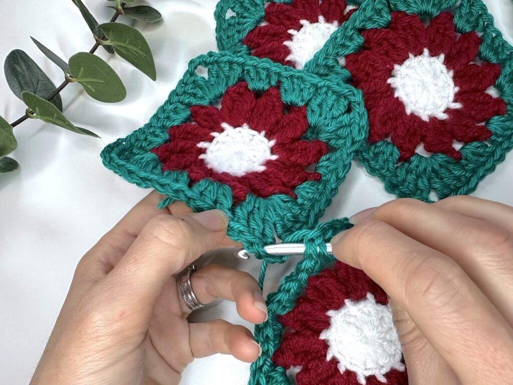 2 hands hold crochet motifs and a hook whilst inserting the hook into stitches on both motifs. The motifs are white, red and green like a flower.