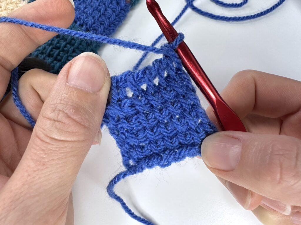 A completed square of Tunisian crochet entrelac in blue before the bind off is done. 2 hands hold up with a red crochet hook