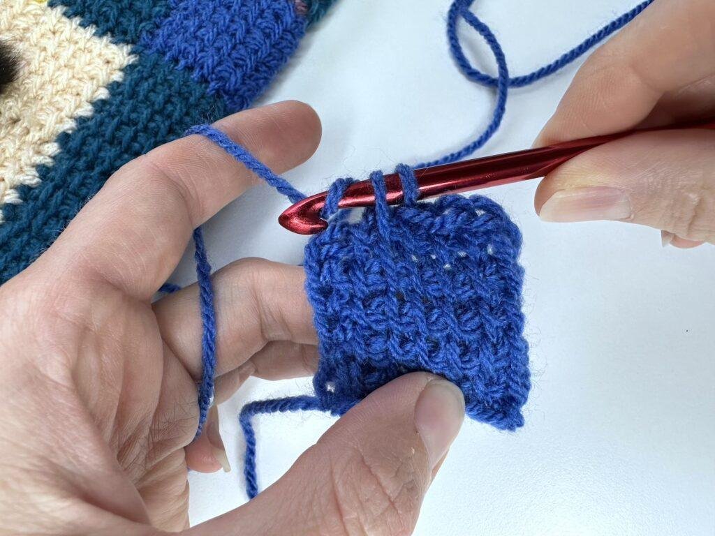 2 hands and a red crochet hook are completing a bind off in Tunisian crochet entrelac for the first square