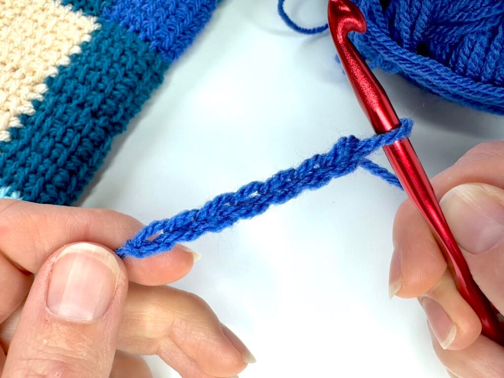 2 hands are holding a chain of crochet in blue and a crochet hook in red as the first step of Tunisian crochet entrelac first square