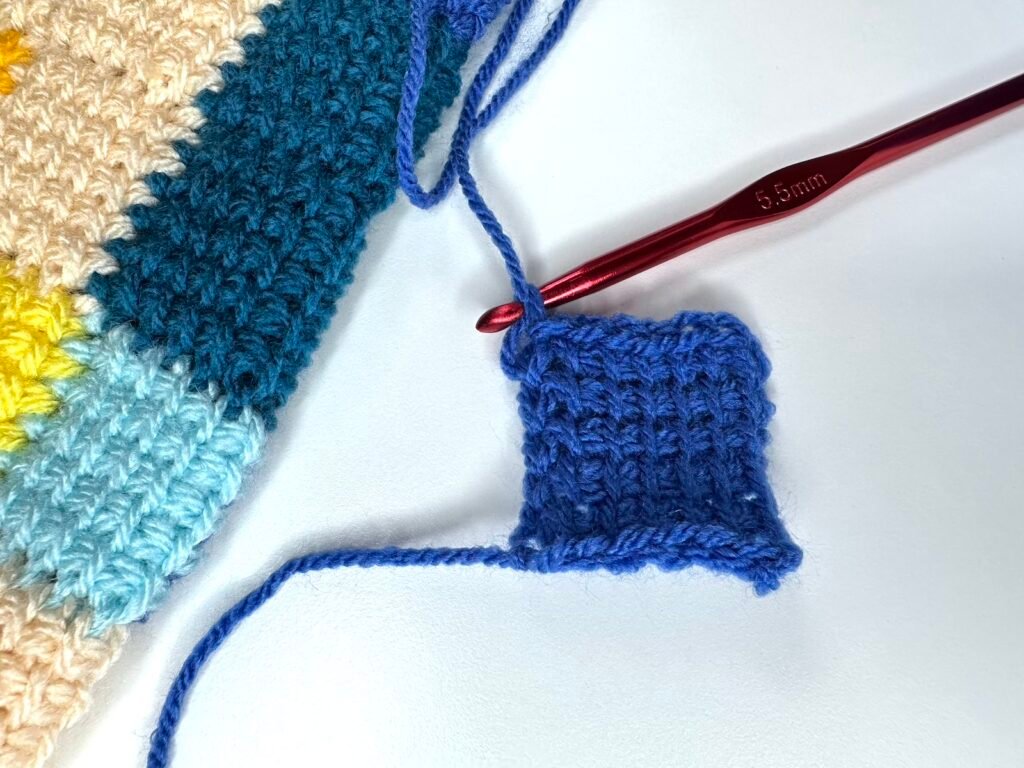 Tunisian crochet entrelac first square in blue with a red crochet hook, and a sample of more Tunisian Crochet entrelac to the left edge.