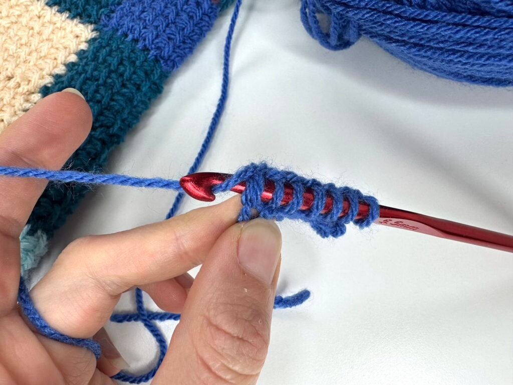 A red crochet hook is being inserted into the end stitch of a Tunisian crochet entrelac first square to show how it is done