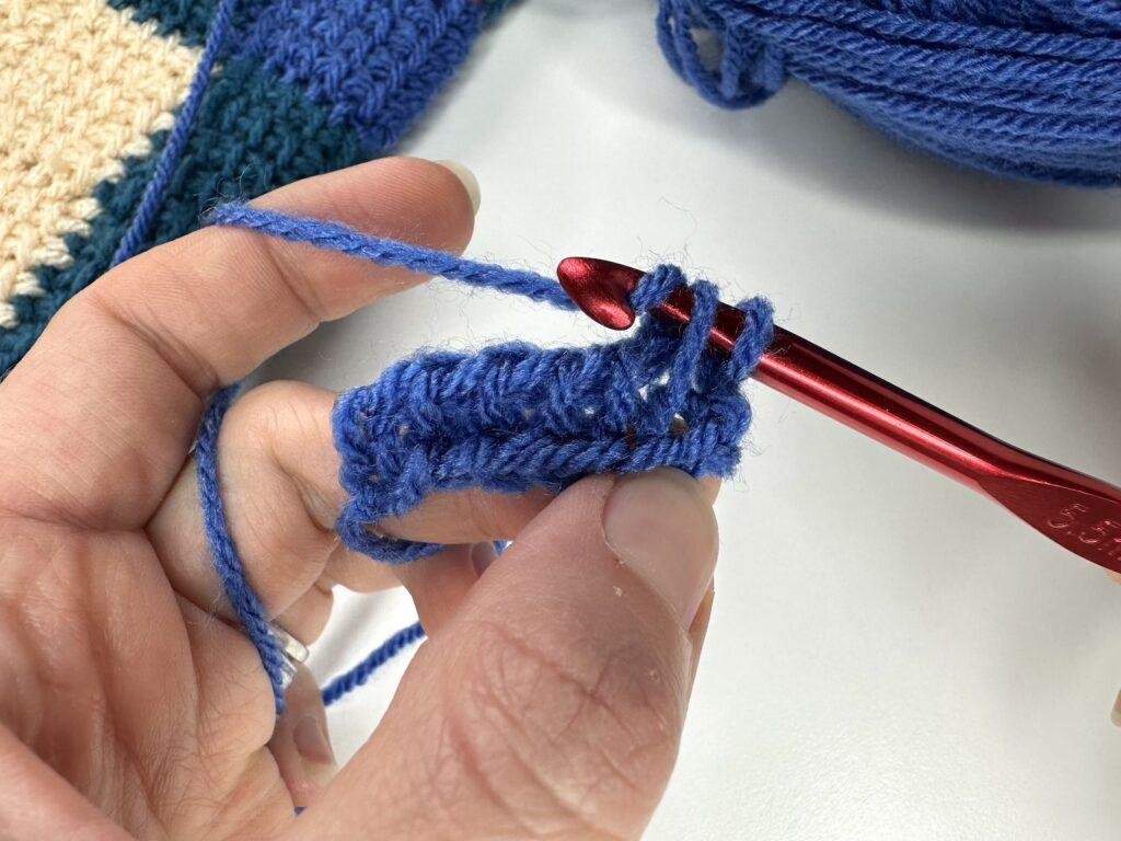 A hand and red crochet hook are showing how to yarn over and pull up a loop in Tunisian crochet entrelac