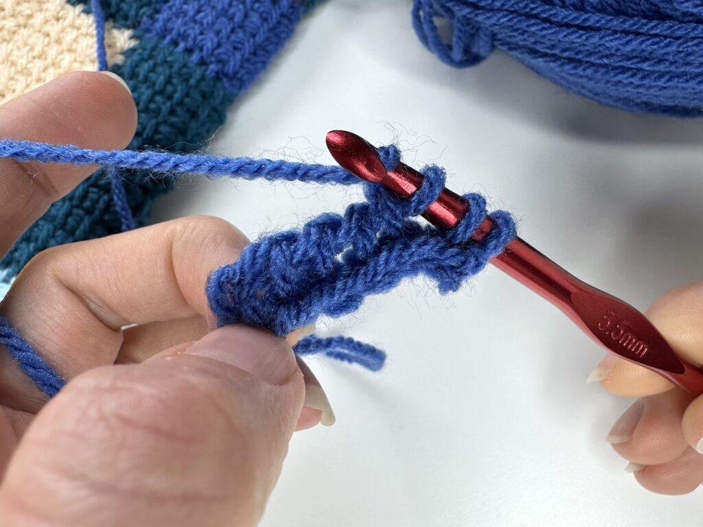 2 hands are showing the Tunisian crochet entrelac first square return pass with blue yarn and a red hook