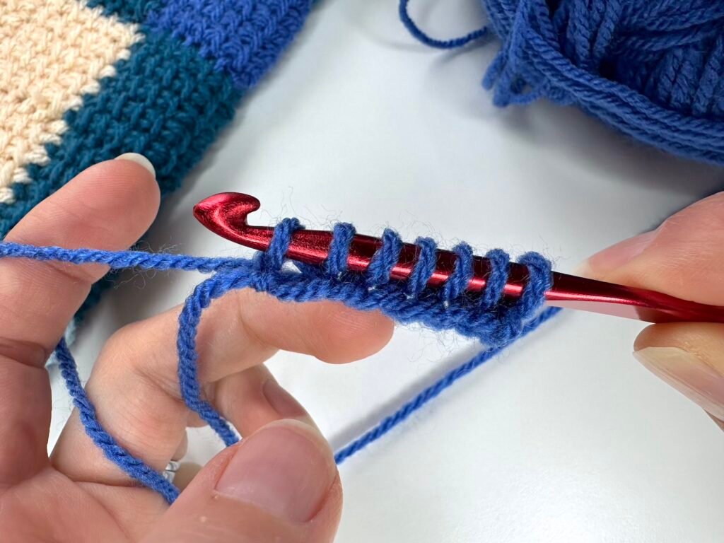 Tunisian crochet entrelac first square set up row is shown with blue yarn and a red hook held in 2 hands. There are 7 loops on the hook now.