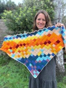 A woman stands in a garden holding up a crochet shawl with lots of different coloured squares all over it
