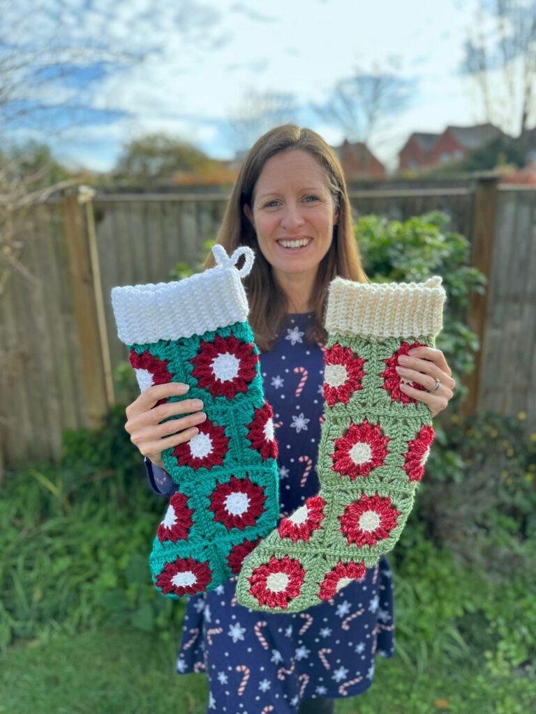 A lady in a garden wearing a blue dress holds 2 crochet poinsettia stockings in front of her