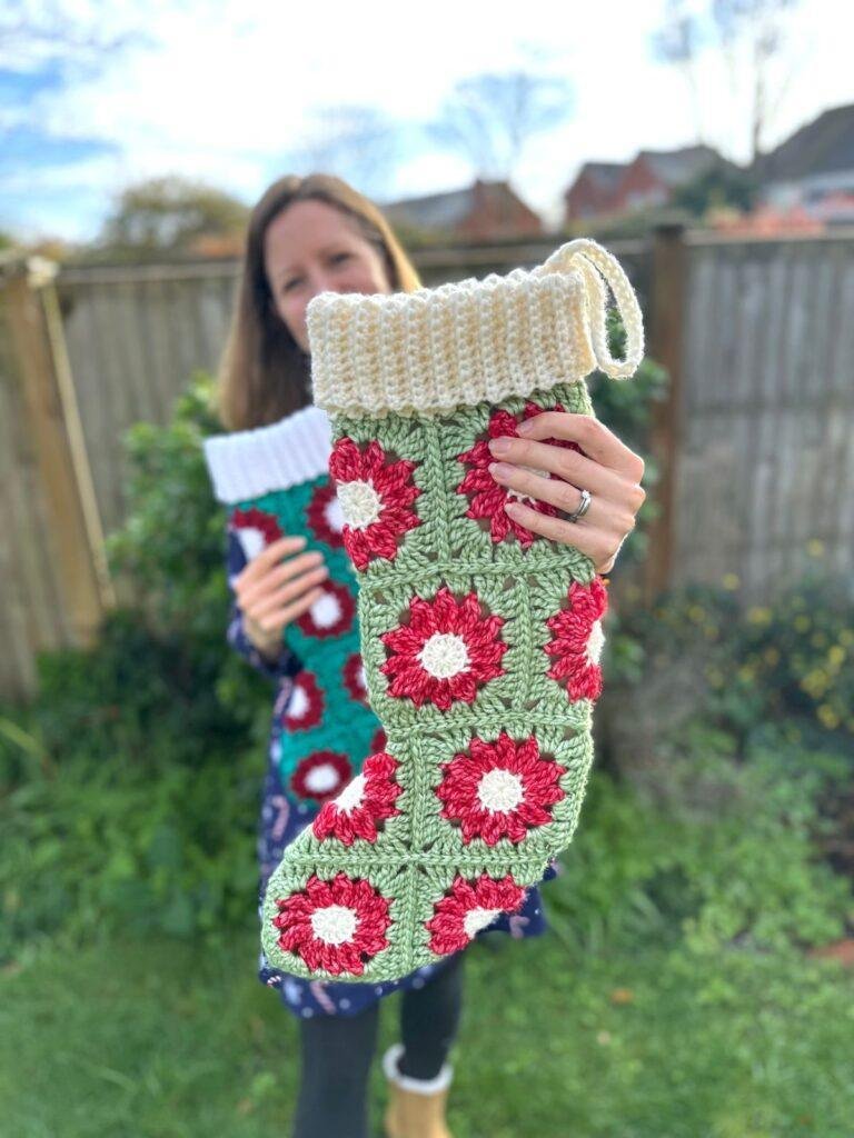 A woman in a garden is holding 2 crochet stockings, holding one out in front to show off the red, white and green granny square flower motifs