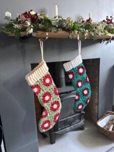 2 crochet stockings made of granny squares that look like red poinsettias or a green background hang on a mantelpiece