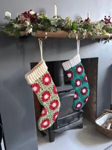 2 crochet stockings made of granny squares that look like red poinsettias or a green background hang on a mantelpiece