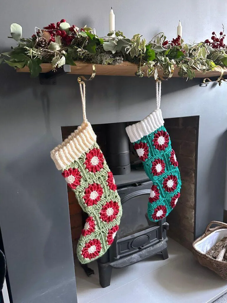 2 crochet stockings made of granny squares that look like red poinsettias or a green background hang on a mantelpiece