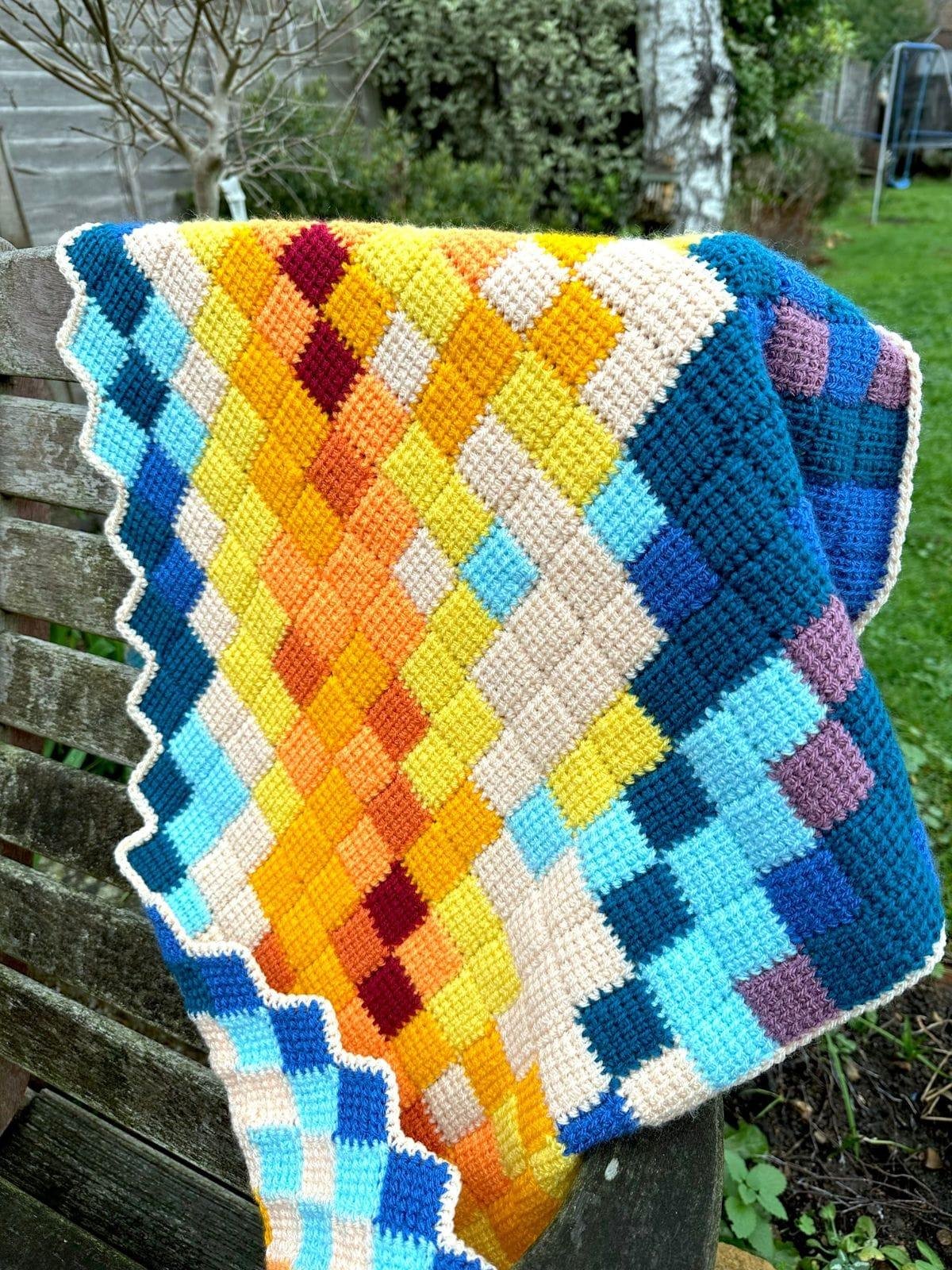 A brightly coloured shawl made out of lots of small crochet squares hangs on a wooden chair in a garden