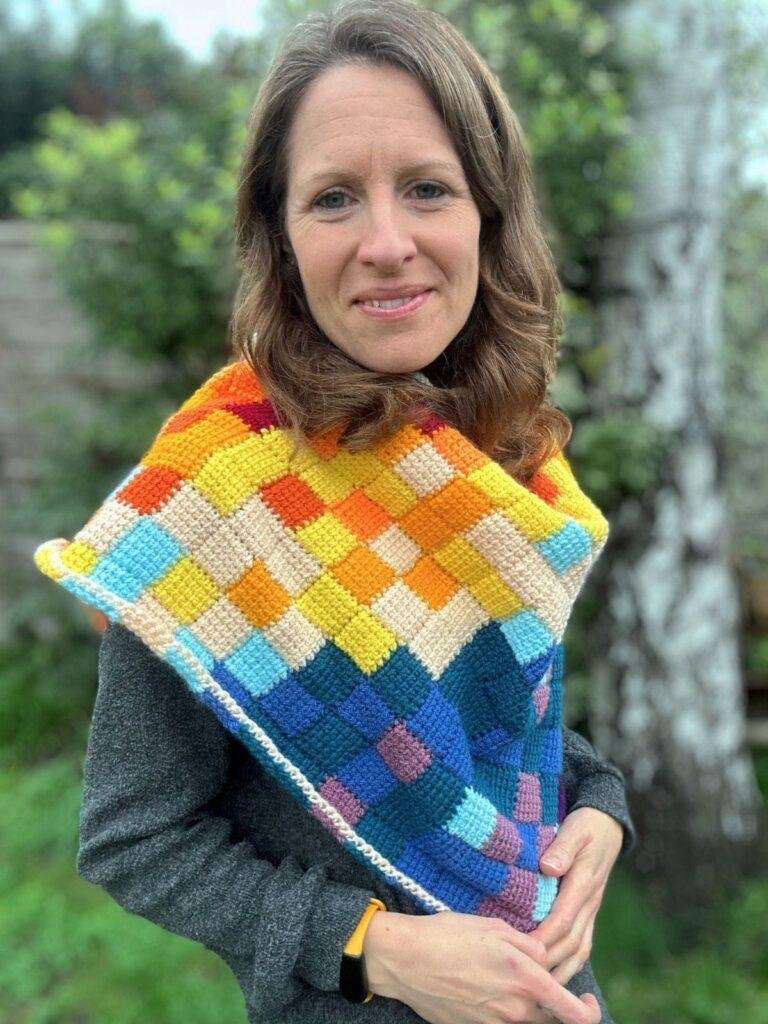 Close up of a lady with brown hair wearing a crochet traingular shawl and standing in a garden. The shawl has lots of squares in different colours to represent different temperatures