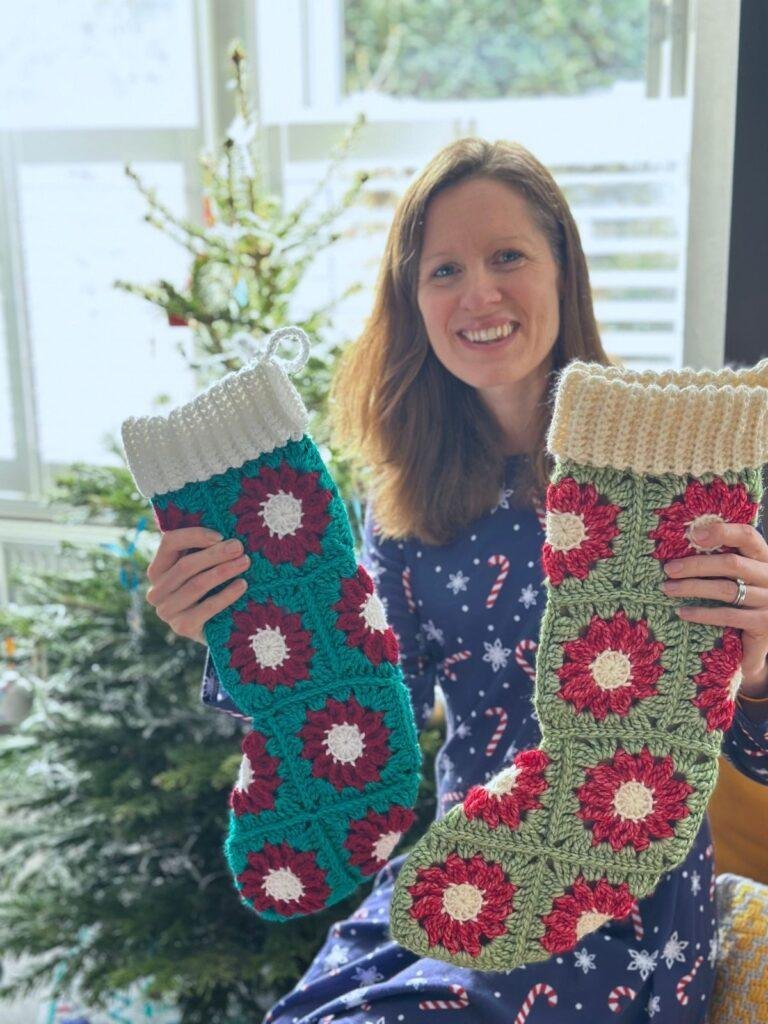A lady smiles and holds 2 crochet stockings in white, red and green, but different tones. She sits in front of a Christmas tree