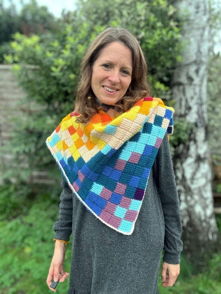 A lady stands in a garden with her head on one side wearing a grey long sleeved dress and a checked, multi-coloured crochet shawl in a triangle shape around her neck