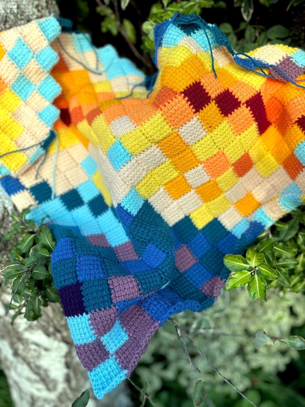 A crochet entrelac shawl is draped in a tree. It is made up of different coloured squares