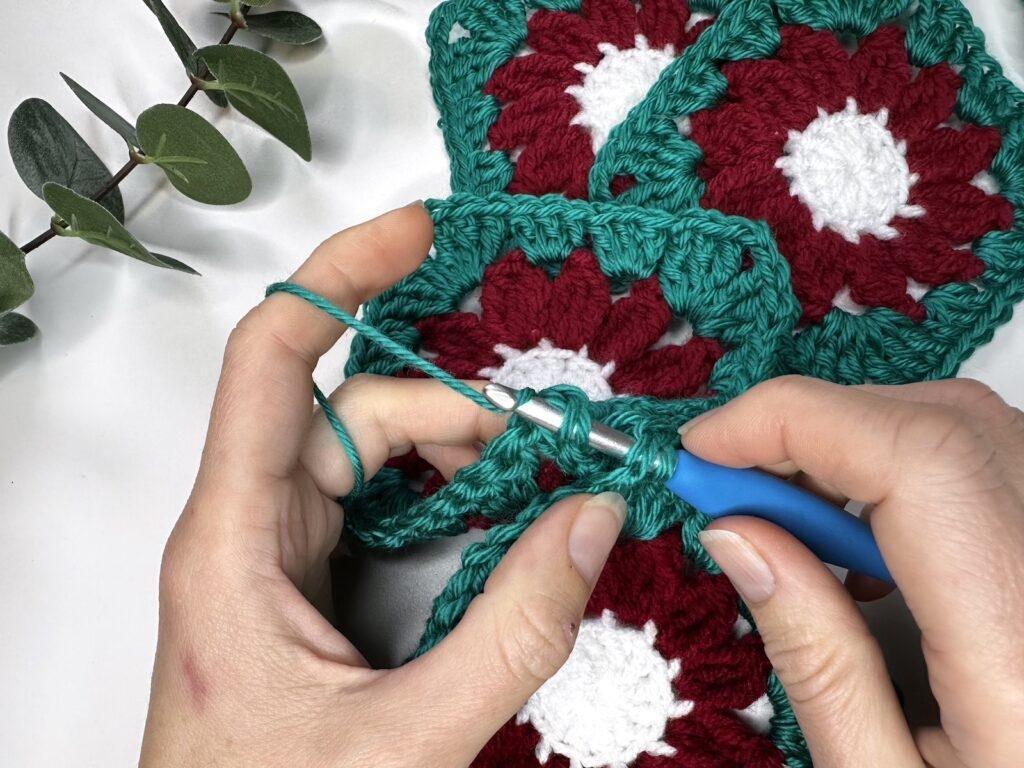2 hands are holding crochet motifs and a crochet hook making a slip stitch by wrapping the yarn around the hook