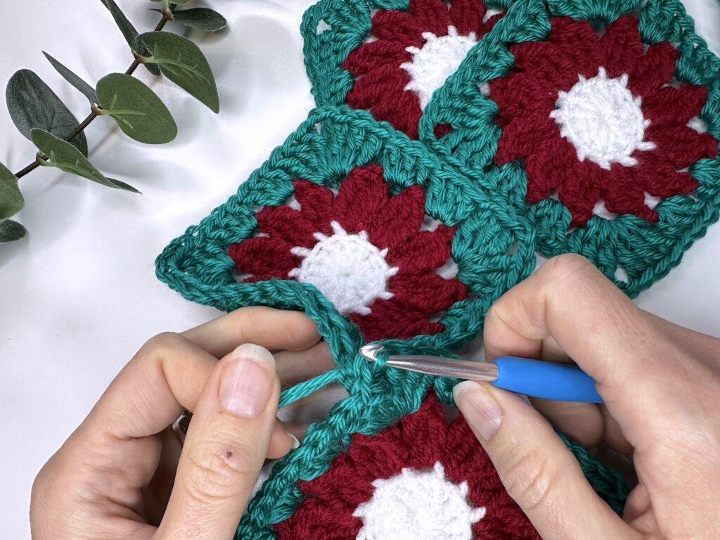 4 flower crochet granny squares in green, red and white, with 2 hands and a crochet hook completing a stitch