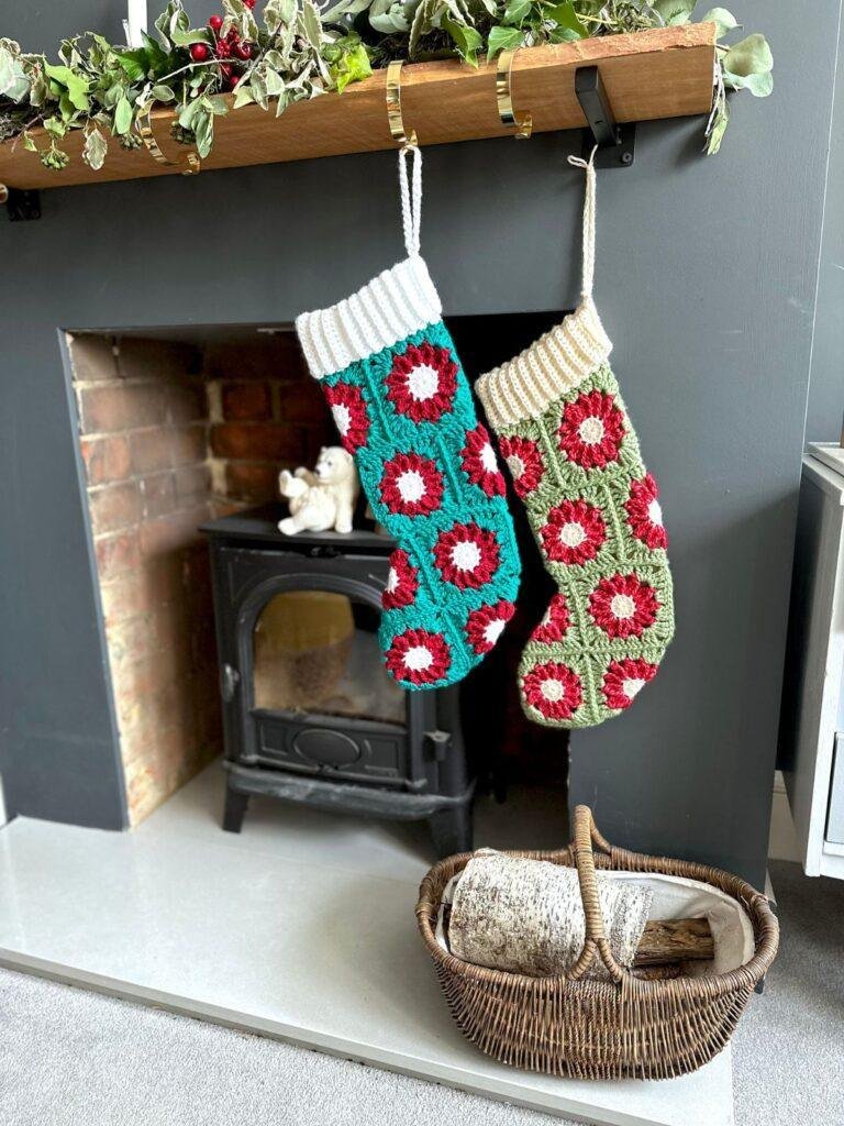 2 crochet granny square stockings hang next to each other on a fireplace with a garland on the mantel and a basket of wood below