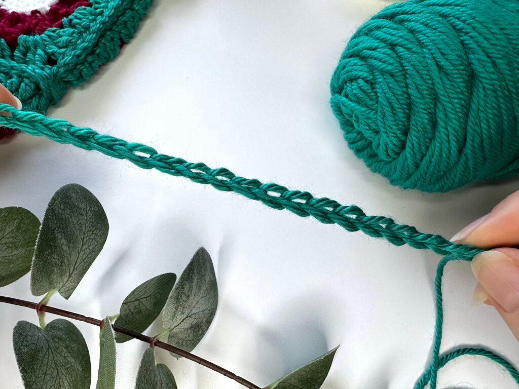 2 hands hold a crochet chain stretched out in green yarn. The ball of yarn is to the side with some greenery in front