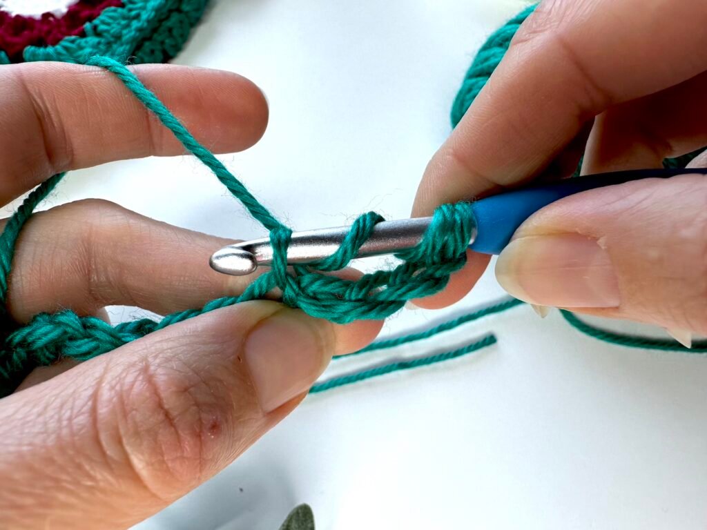 Midway through a crochet stitch, 2 hands with hook and green crochet yarn insert a hook into the stitch