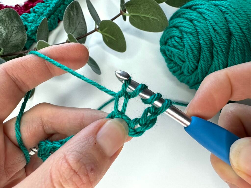 Partway through a treble crochet stitch, 2 hands are holding green yarn with 4 loops showing on the crochet hook