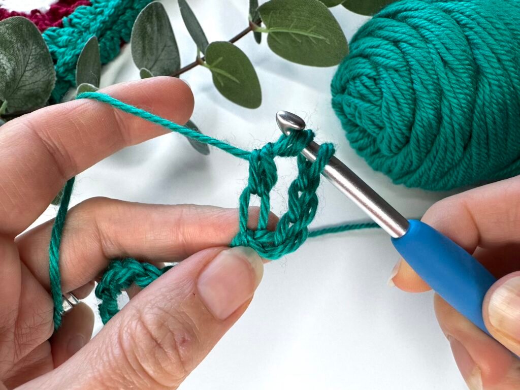 2 hands are holding a crochet hook and yarn with a partly completed treble crochet stitch on the hook