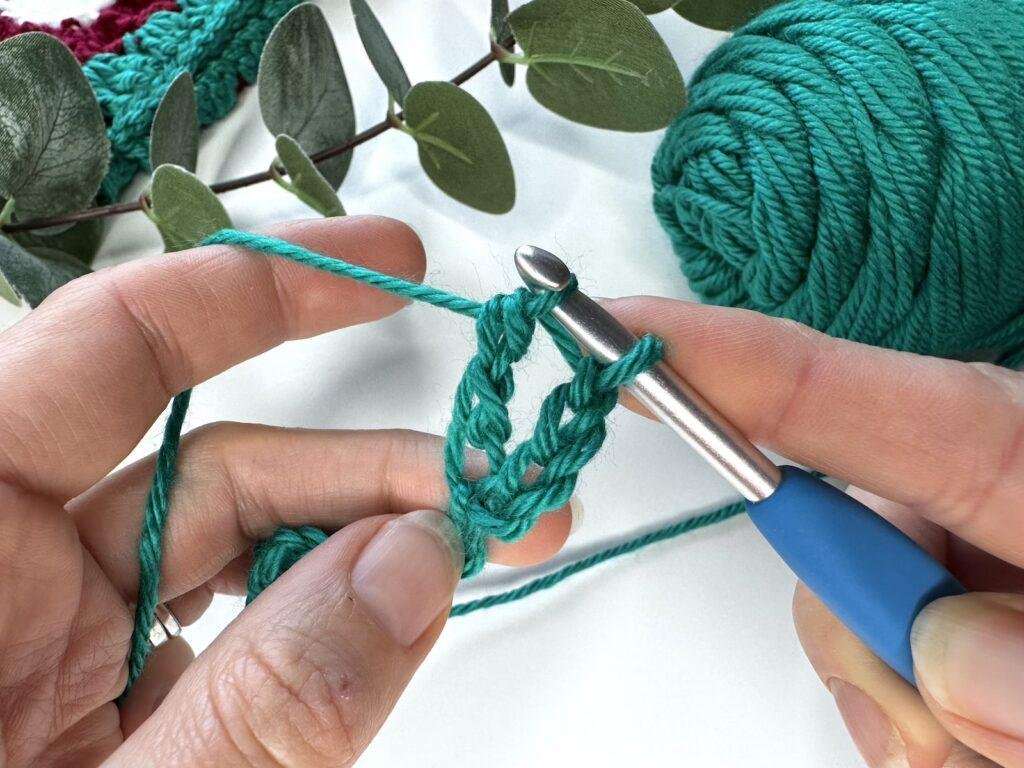 2 hands hold green yarn and crochet hook and have almost finished a triple crochet stitch