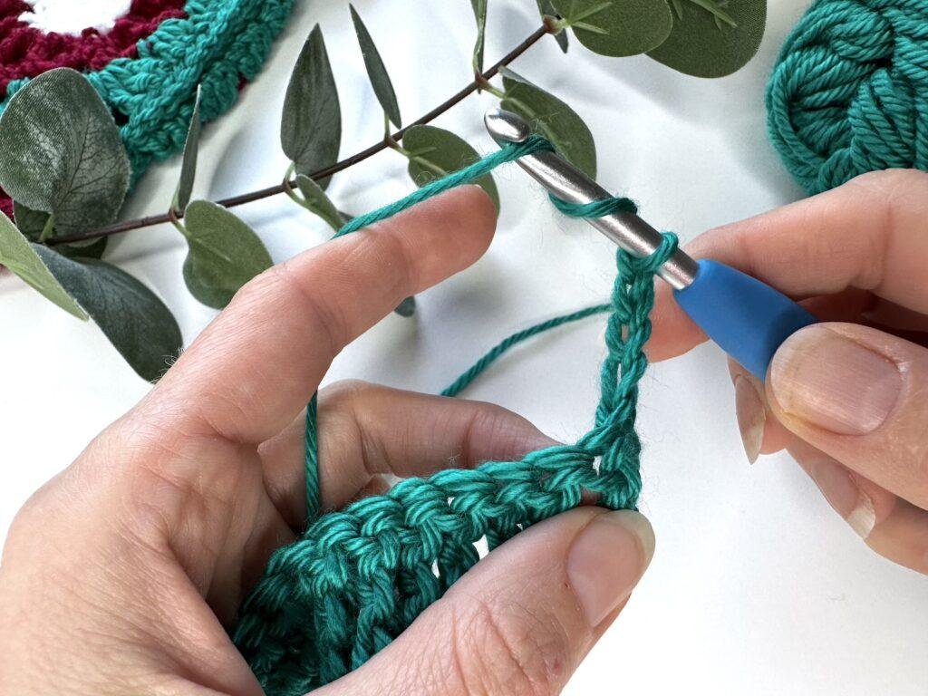 2 hands are moving onto a second row on crocheting using green yarn and a blue crochet hook