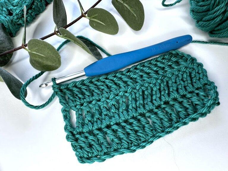 A swatch of green treble crochet stitches with a hook joined on the top and some greenery above