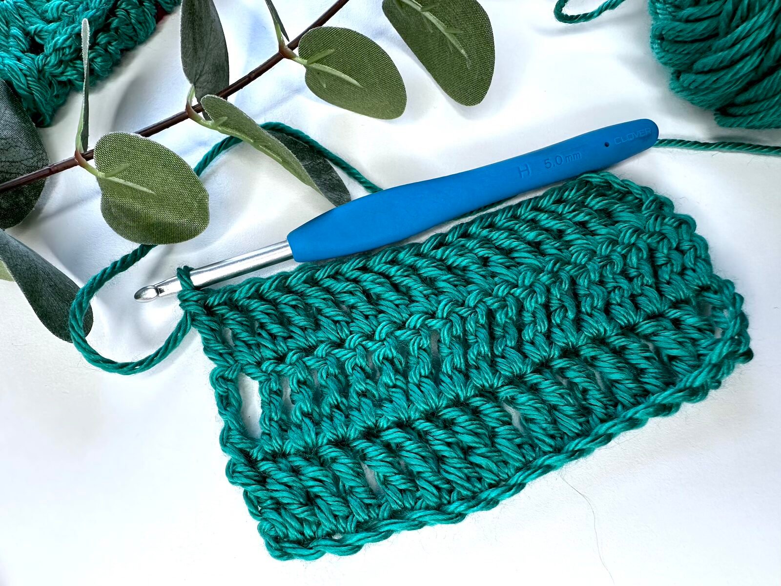 A swatch of green treble crochet stitches with a hook joined on the top and some greenery above