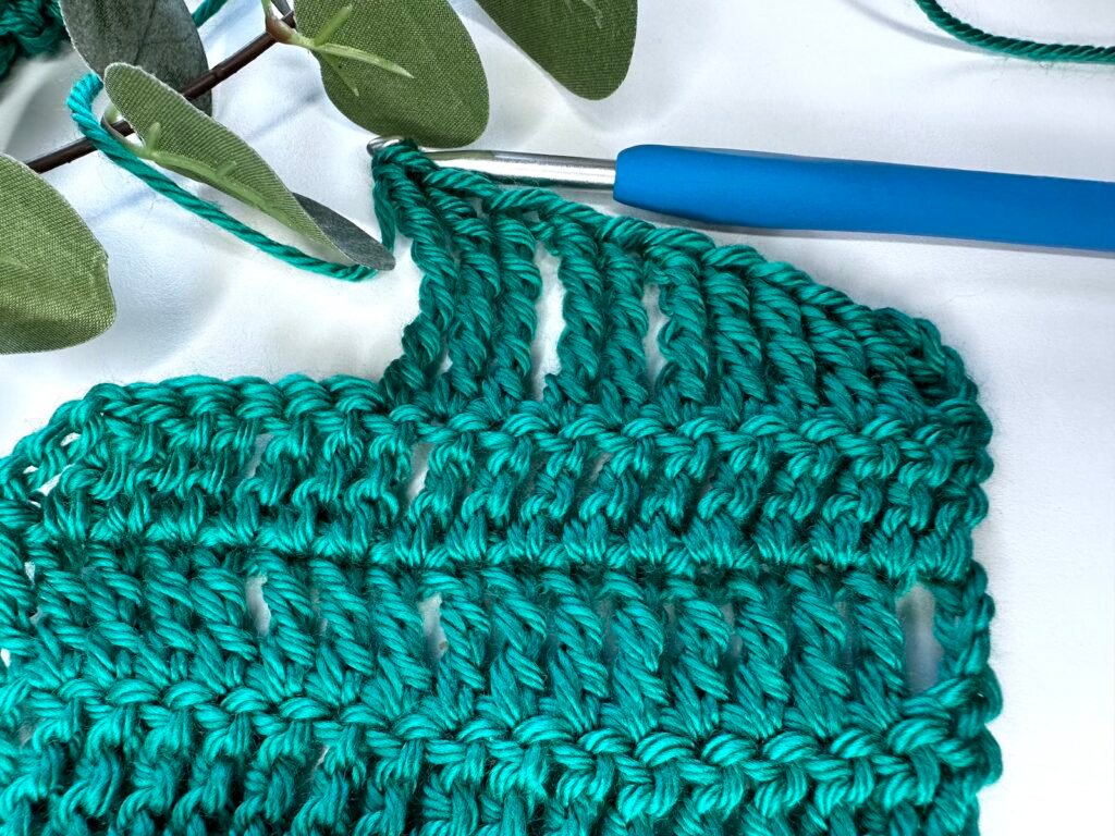A picture of a green swatch of treble crochet stitches and then at the top there are even taller crochet stitches getting taller across the row. The hook is still joined on and there is greenery above