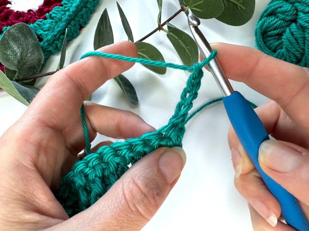 A green crochet fabric is being made and a chain has just been created by 2 hands and a blue crochet hook