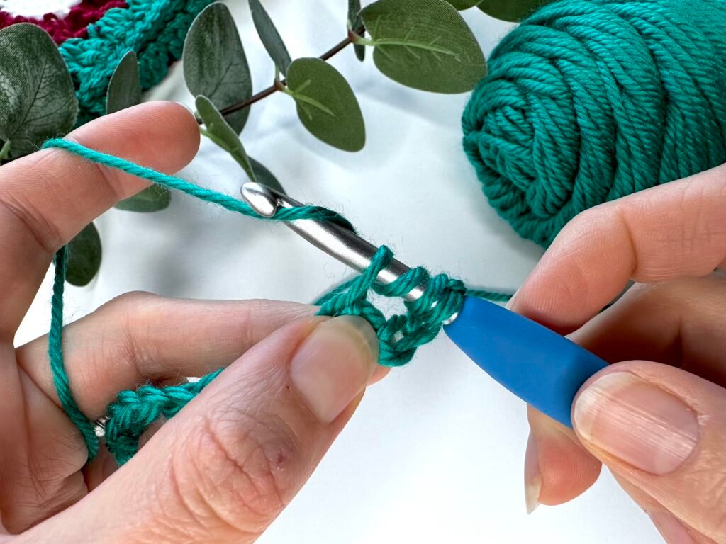 2 hands with green crochet fabric are making a stitch by wrapping the yarn around a crochet hook